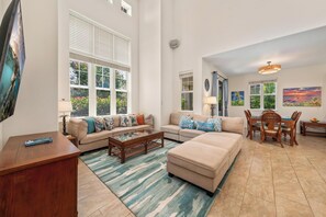 A spacious living room with a large window, a dining area with a table and chairs, and a cozy seating area with a sofa and ottoman. - 140” Movie Projector and Beach Gear Included. 2201 Waikoloa Colony Villas (Waikoloa Beach Resort)