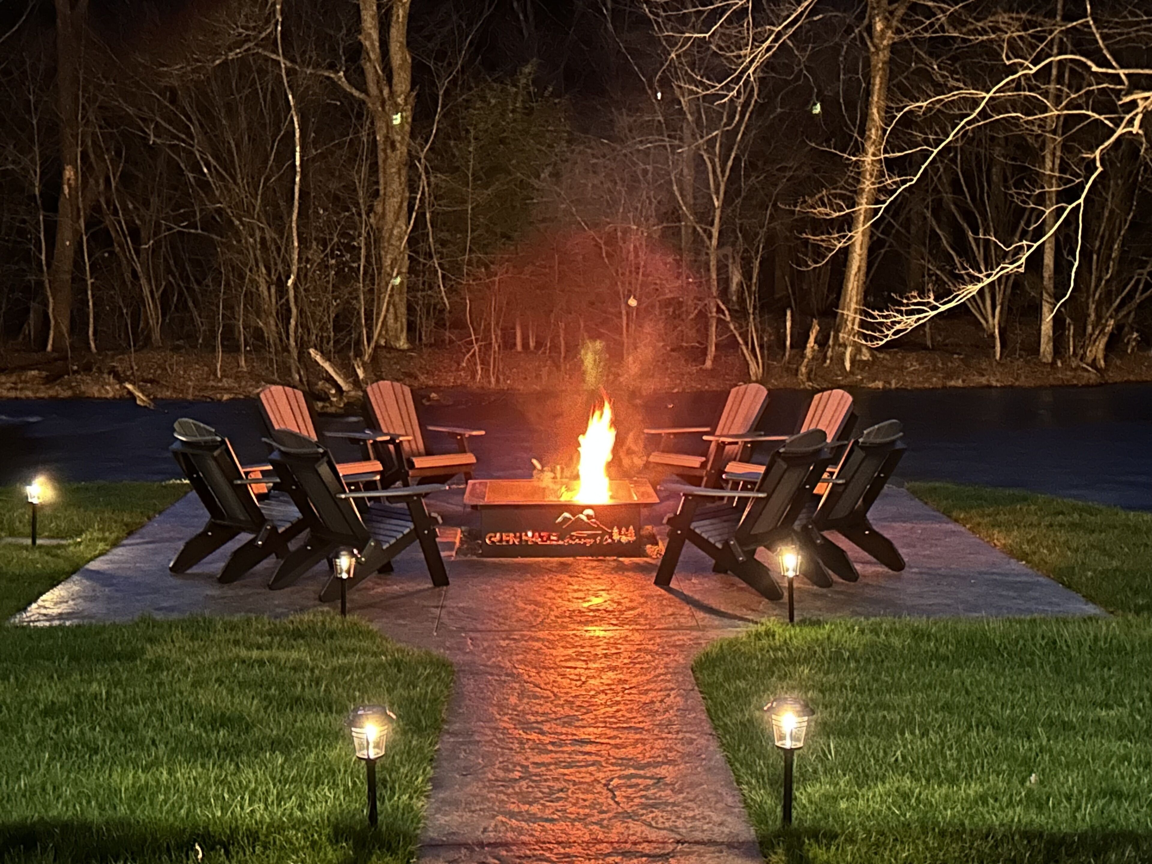 Un hogar de fuego rodeado de sillas Adirondack en un patio de concreto, con un sendero que conduce hacia él.