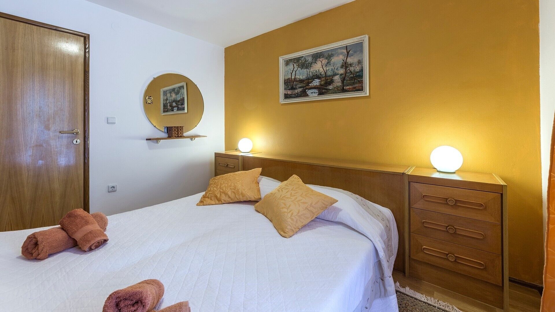 Ein Hotelzimmer mit einem Bett, zwei Nachttischen mit Lampen, einem Holzschrank, einem runden Spiegel und einem Bild an der Wand.