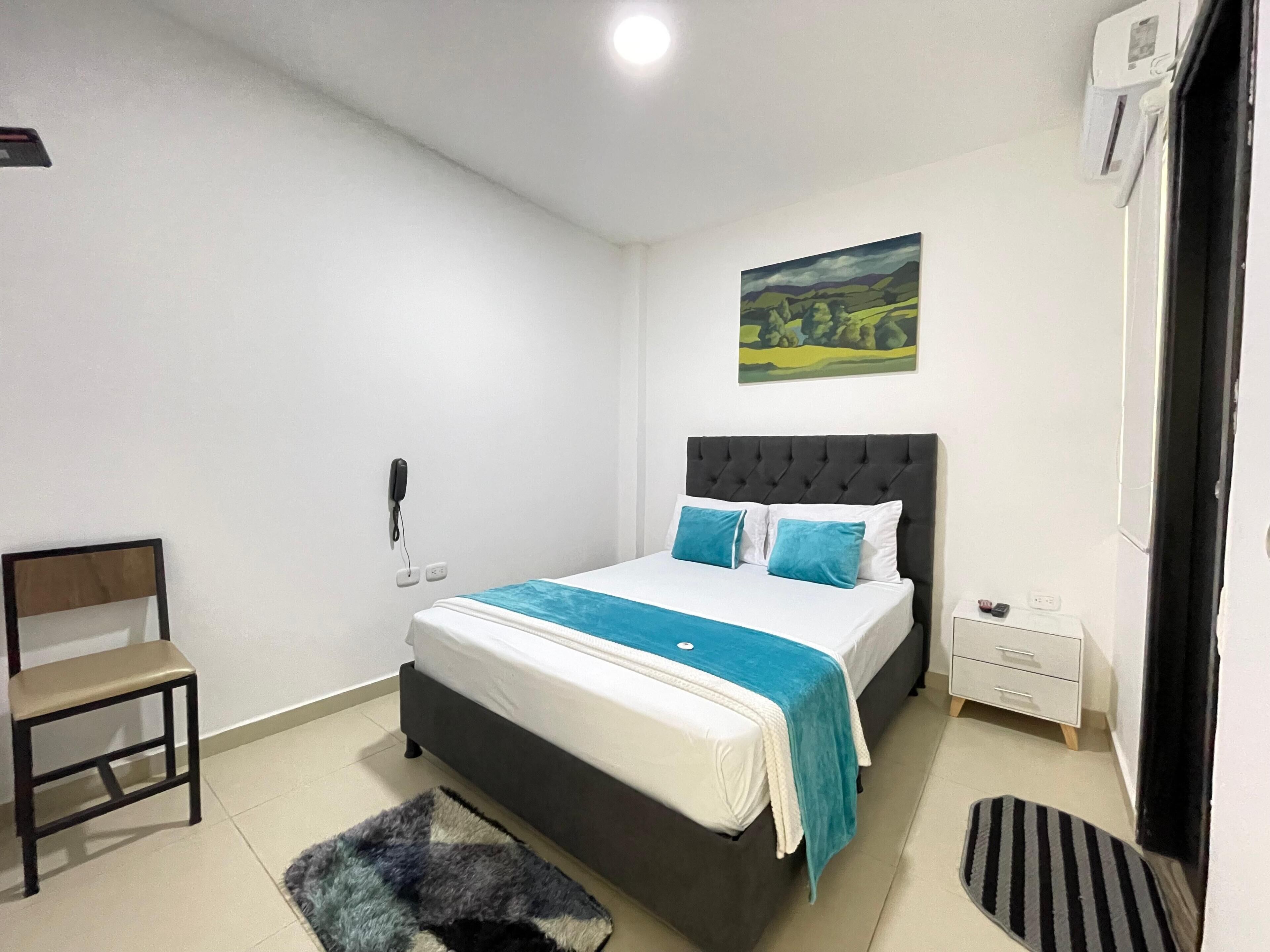 A bedroom with a bed, a chair, a bedside table, a nightstand, a painting, and a door.