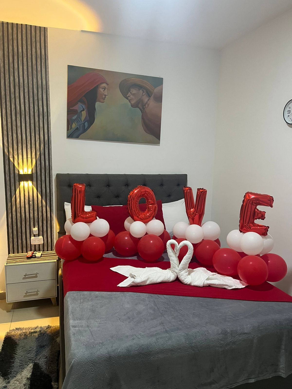 A bedroom with a bed covered in red and white balloons spelling "LOVE," a painting on the wall, and a clock.