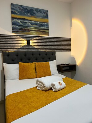 A neatly made bed with a white and mustard-colored bedspread, a folded towel, and a headboard with a wooden panel design. - HOTEL ANAJIRAWAA (Riohacha)