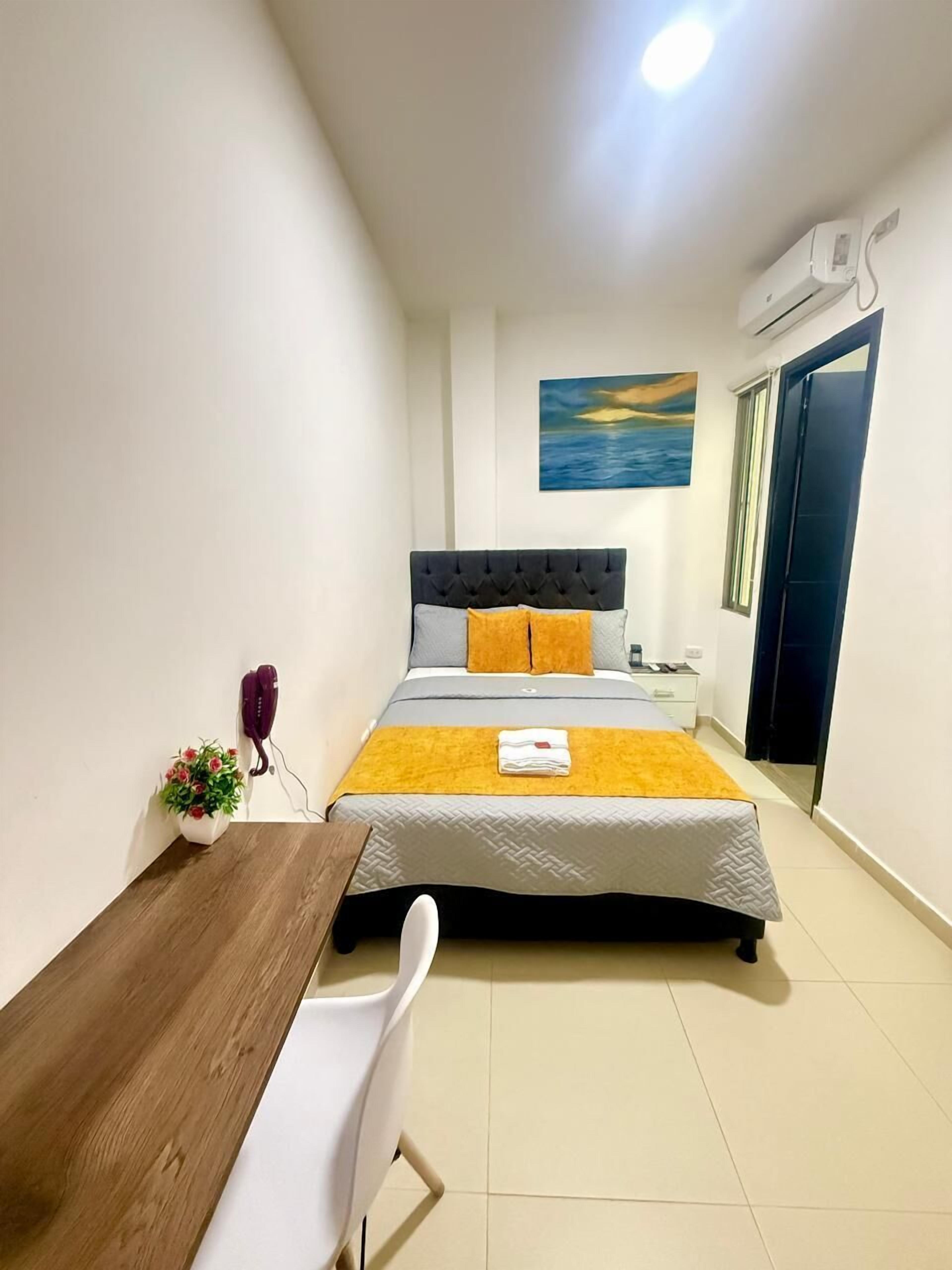 Un dormitorio con cama, mesita de noche, una silla, una pequeña mesa con una planta y un cuadro en la pared.
