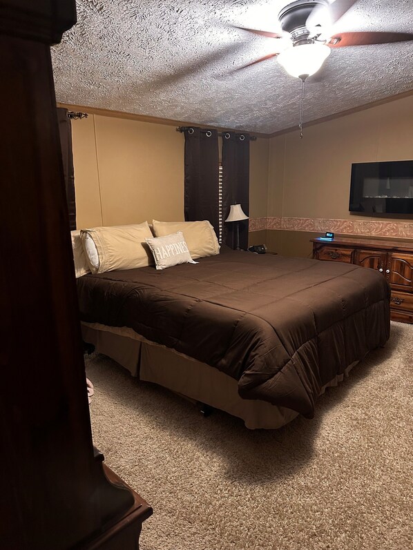 Un dormitorio con una cama grande, un ventilador de techo, un televisor montado en la pared y una mesita de noche con una lámpara.