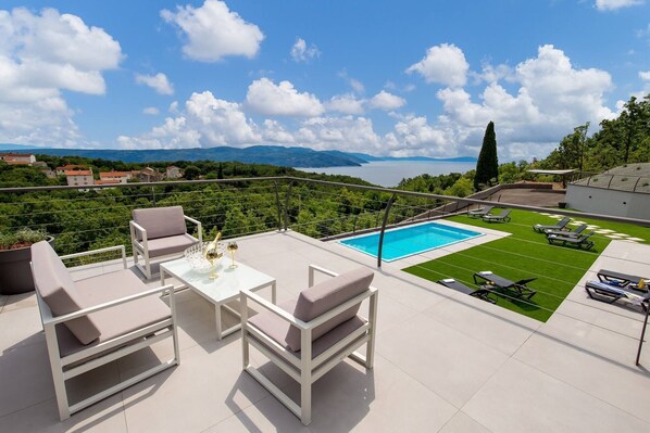 Relax on the spacious outdoor terrace with a clear view of the pool and lush surroundings, surrounded by comfortable lounge chairs and a glass table.
