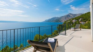 Ein Balkon mit zwei Liegestühlen, einer mit Handtuch, Blick auf das Meer mit Bergen im Hintergrund.