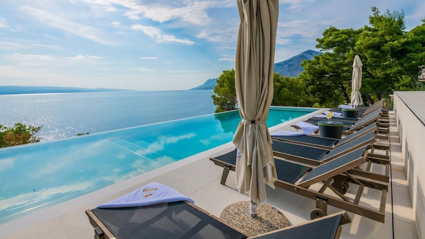 Ein Poolbereich mit Liegestühlen und Sonnenschirmen, mit Blick auf ein klares, blaues Meer und eine gebirgige Landschaft.
