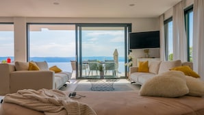 Ein modernes Wohnzimmer mit einem großen Schiebeglastür, die auf einen Balkon mit Meerblick führt. Es gibt ein Sofa, einen Couchtisch und einen an der Wand befestigten Fernseher.