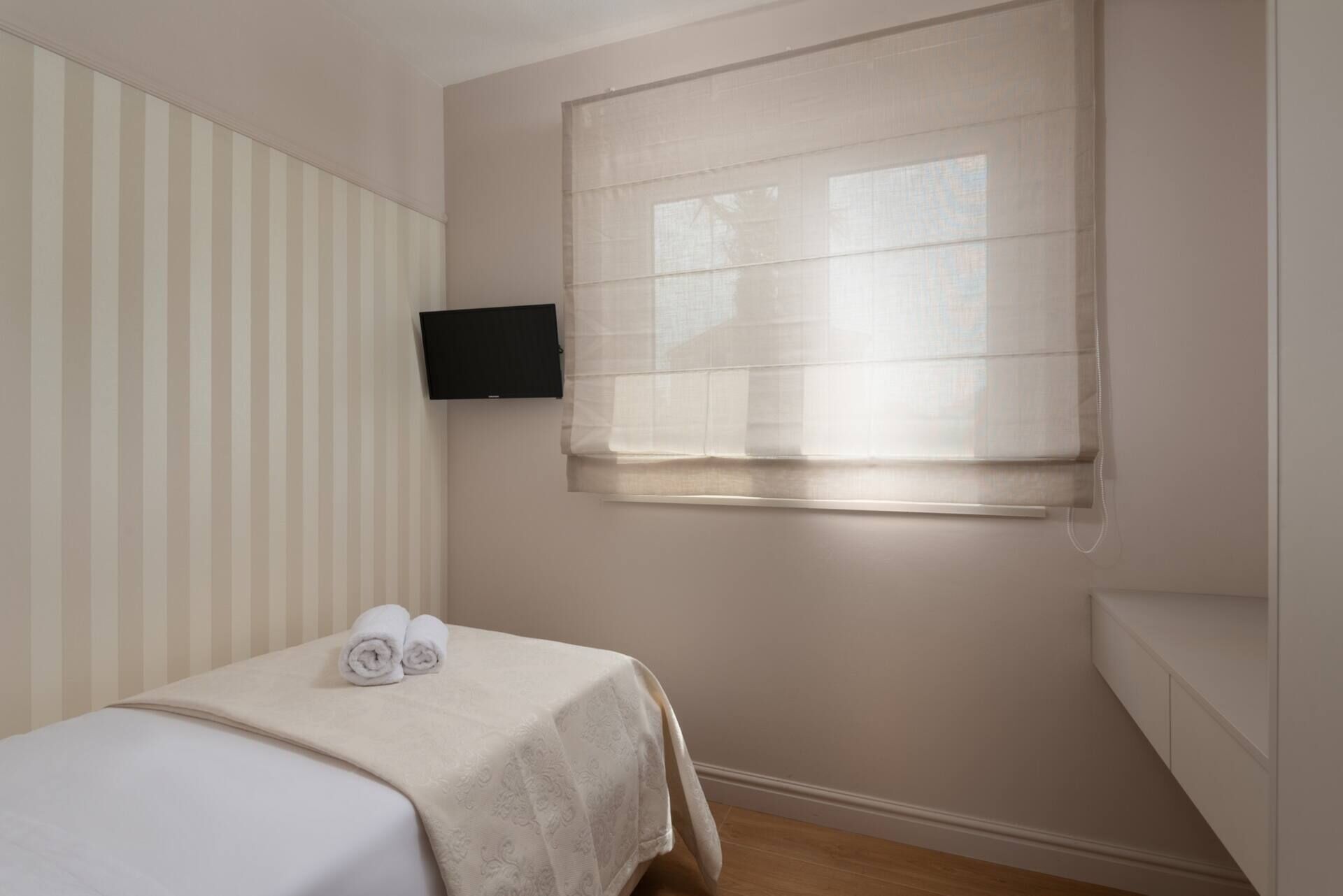 Une petite chambre à coucher, bien rangée, avec un lit, un téléviseur fixé au mur et une fenêtre équipée de stores.