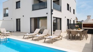 Een modern huis met twee verdiepingen, een zwembad, terrasmeubels en een barbecueplek.
