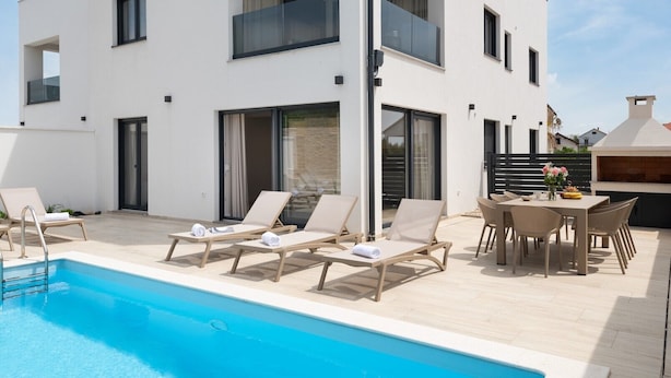 A modern two-story building with a swimming pool, lounge chairs, and a barbecue area.