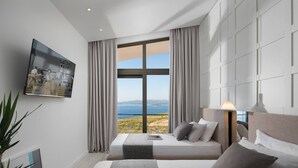 Una habitación de hotel moderna con un amplio ventanal que ofrece vistas panorámicas, una cama con ropa de cama gris y un televisor montado en la pared.