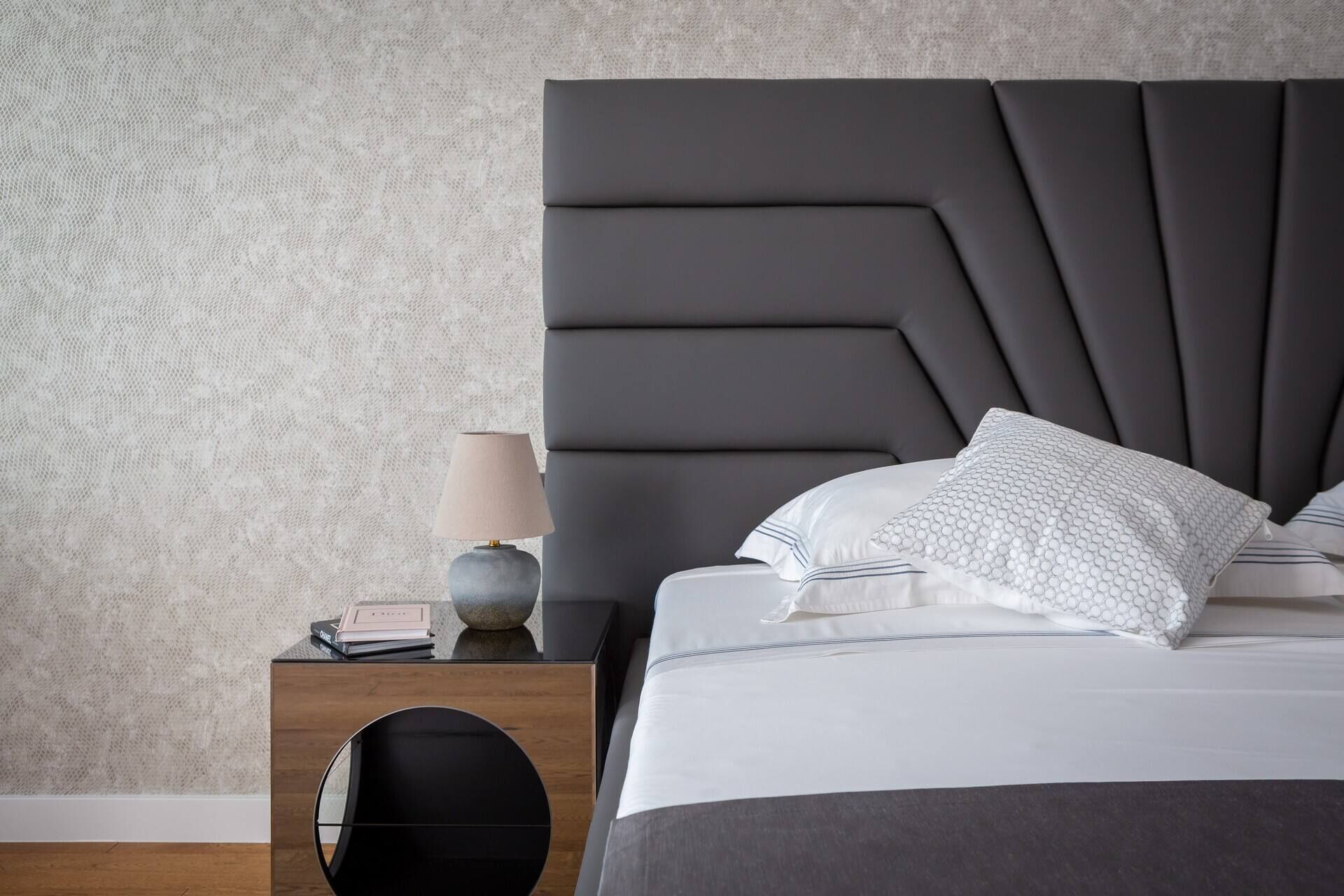 Quarto moderno com cabeceira escura, roupa de cama branca e um espelho redondo sobre uma mesa de cabeceira de madeira.