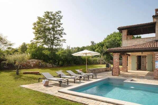A swimming pool with lounge chairs and an umbrella, surrounded by a stone wall and greenery.