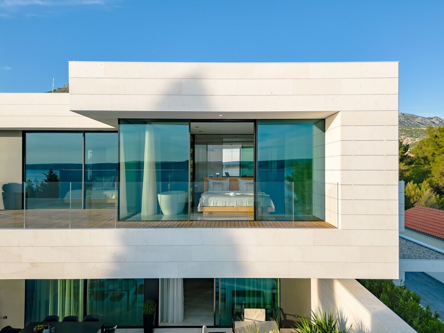 A modern two-story house with large glass windows, a balcony with a wooden deck, and a bedroom with a bed and a bathtub.