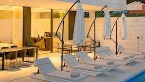 A modern two-story building with a swimming pool, lounge chairs, and umbrellas.