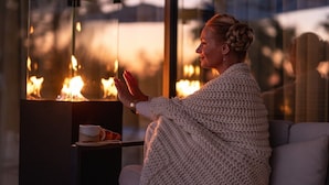 A person sitting by a fireplace with a glass enclosure, a cup, and a plate on a small table beside them.