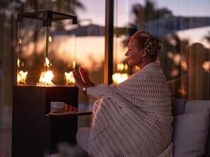A person sitting by a fireplace with a glass enclosure, a cup, and a plate on a small table beside them.