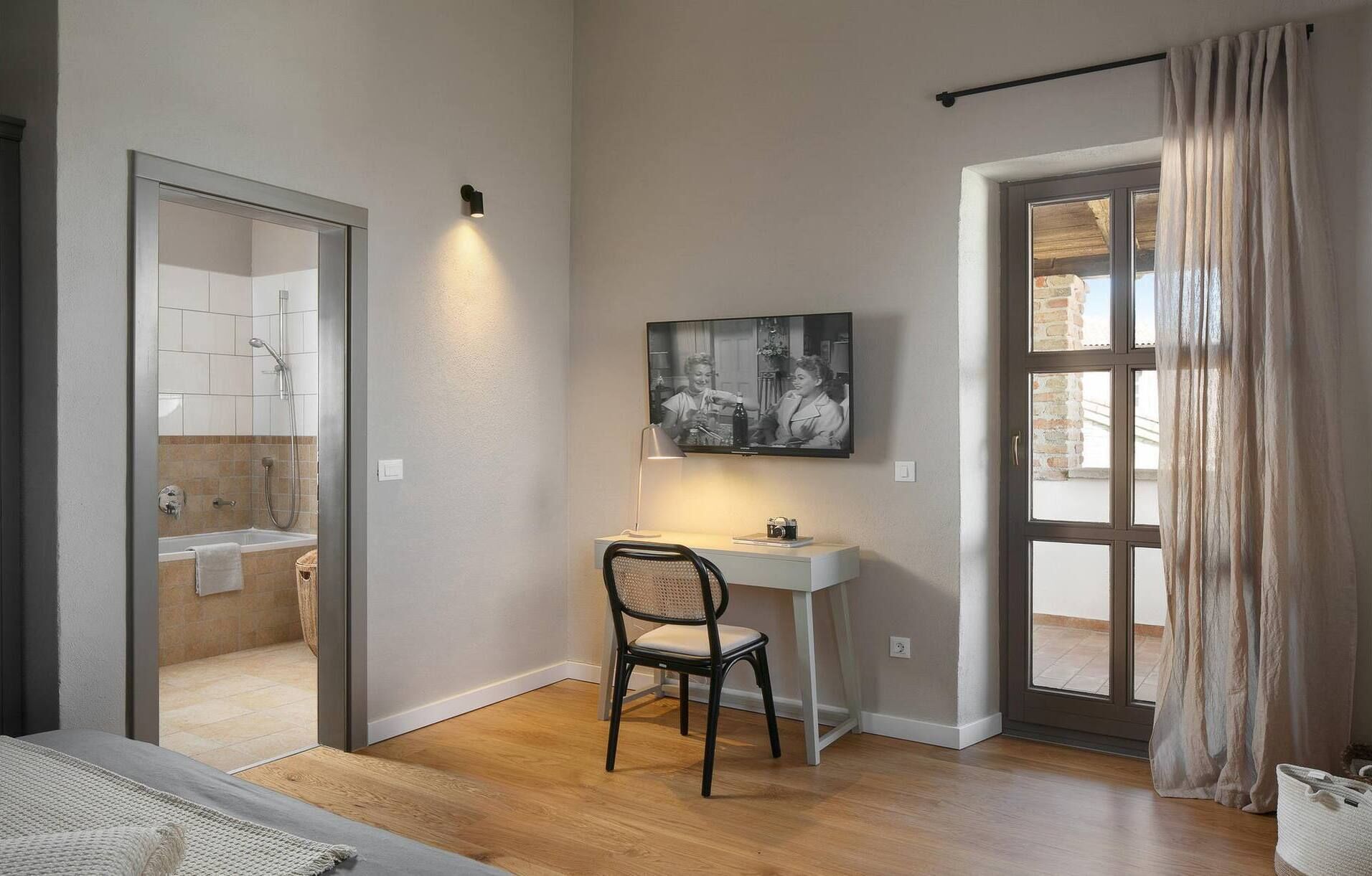 A room with a shower, a desk with a chair, a television, and a large window with curtains.