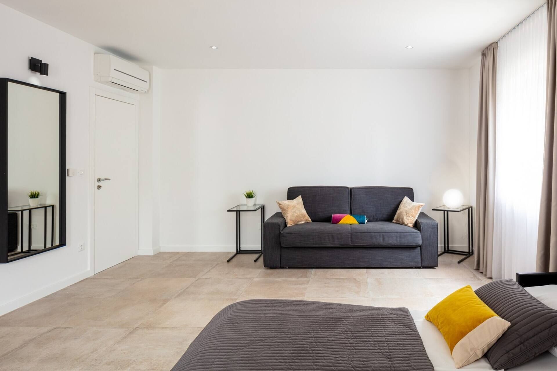 Une chambre à coucher moderne et minimaliste, avec un canapé gris, un lit recouvert d’une couette grise et jaune, et une petite table de chevet avec une lampe.