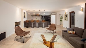 A modern living room with a glass coffee table, two brown chairs, and a sofa with a cushion. A kitchen with a bar area and pendant lighting is visible in the background. - Villa Clementine with pool, sea view, big group near sea pet friendly bbq family (Sutivan)