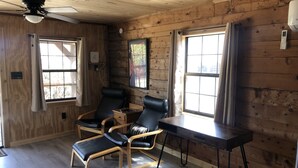 Une pièce aux murs en bois et dotée d’un ventilateur de plafond, comprenant deux fauteuils en cuir noir, une table en bois et de grandes fenêtres avec des rideaux.