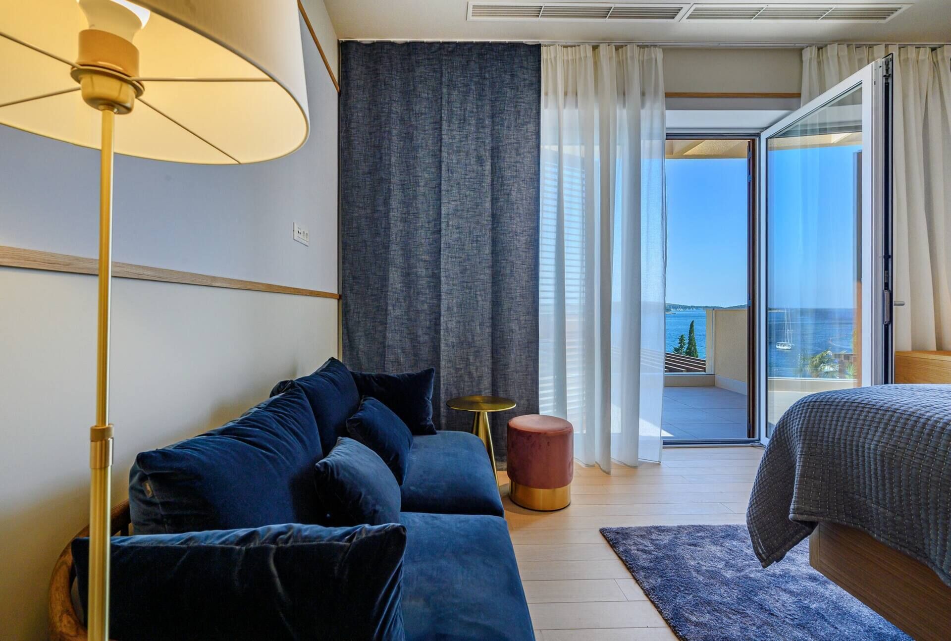 Un dormitorio moderno con un sofá azul, una mesita redonda, una cama y vistas al mar a través de amplios ventanales.