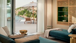 Ett modernt hotellrum med ett stort fönster med utsikt över en pool och en vacker omgivning. Rummet har en turkosfärgad soffa, ett litet runt bord och en bokhylla med dekorativa föremål.