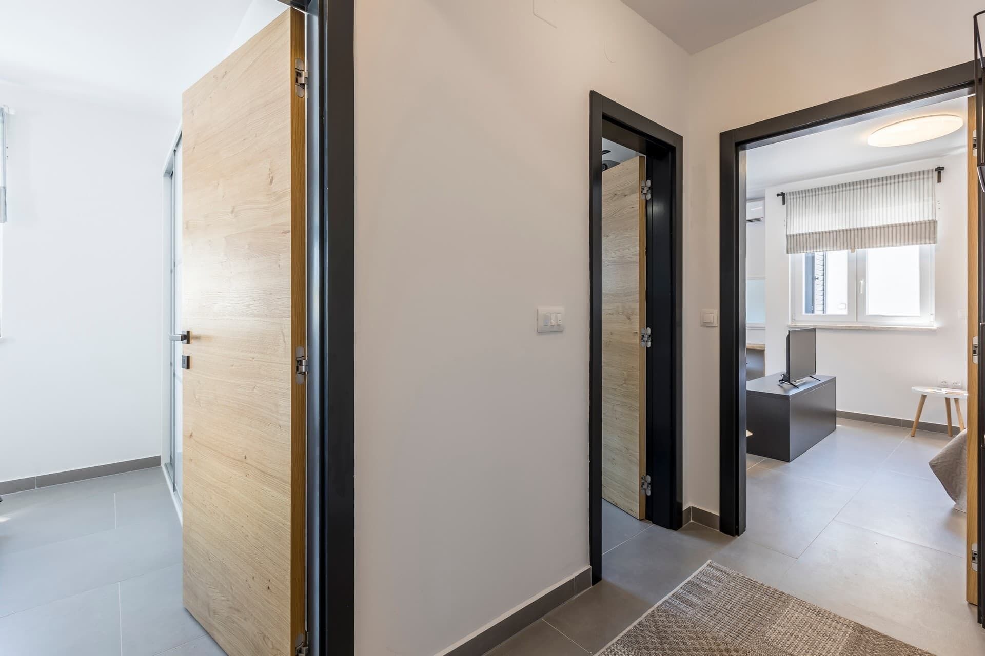 Un couloir avec une porte en bois, un miroir à cadre noir et une fenêtre avec des stores.