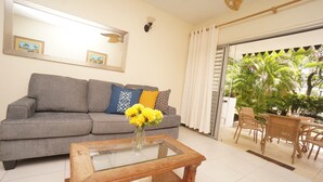 Una sala de estar con un sofá gris, una mesa de centro de vidrio con un jarrón de flores amarillas, y vistas a un patio exterior con mobiliario de mimbre y plantas.