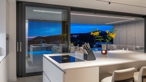 A modern kitchen with a large island, a glass door leading to a balcony, and a view of a cityscape at dusk.
