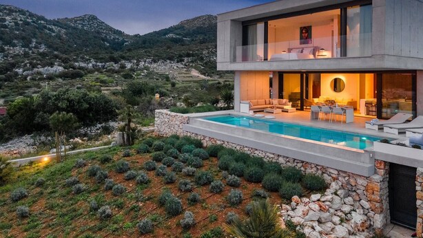 A modern two-story house with a swimming pool, stone walls, and a view of the surrounding landscape.