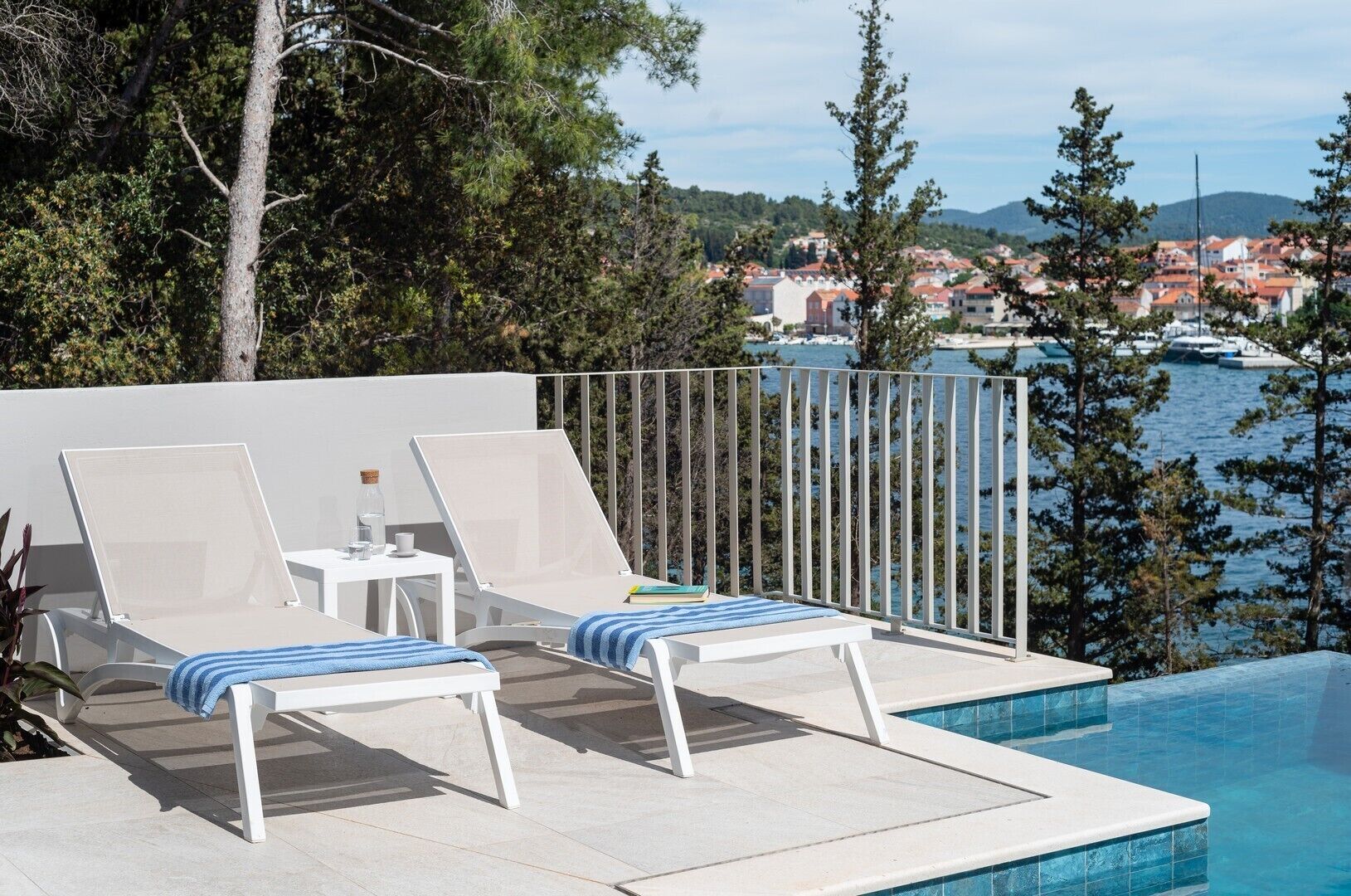 A poolside area with two lounge chairs and a small table, overlooking a marina and a town.