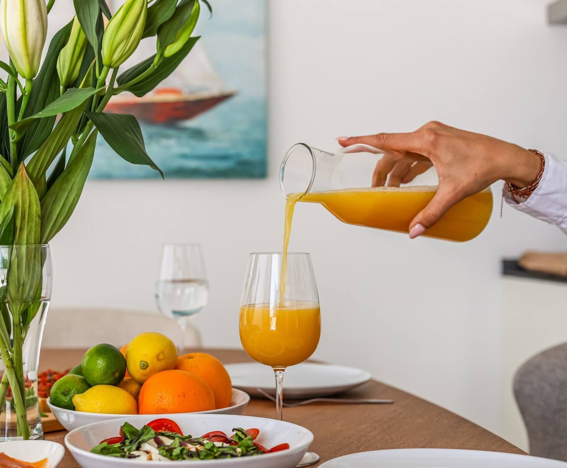 Een hand giet sinaasappelsap in een glas. Op de tafel staan fruit en een salade.
