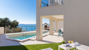 Un espace piscine extérieur moderne offrant une vue dégagée sur la mer, un coin barbecue et un espace détente avec une table et des chaises.