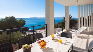Un balcon avec une table dressée pour le petit-déjeuner, agrémentée de fruits et de jus, offrant une vue panoramique sur la mer et les montagnes lointaines.