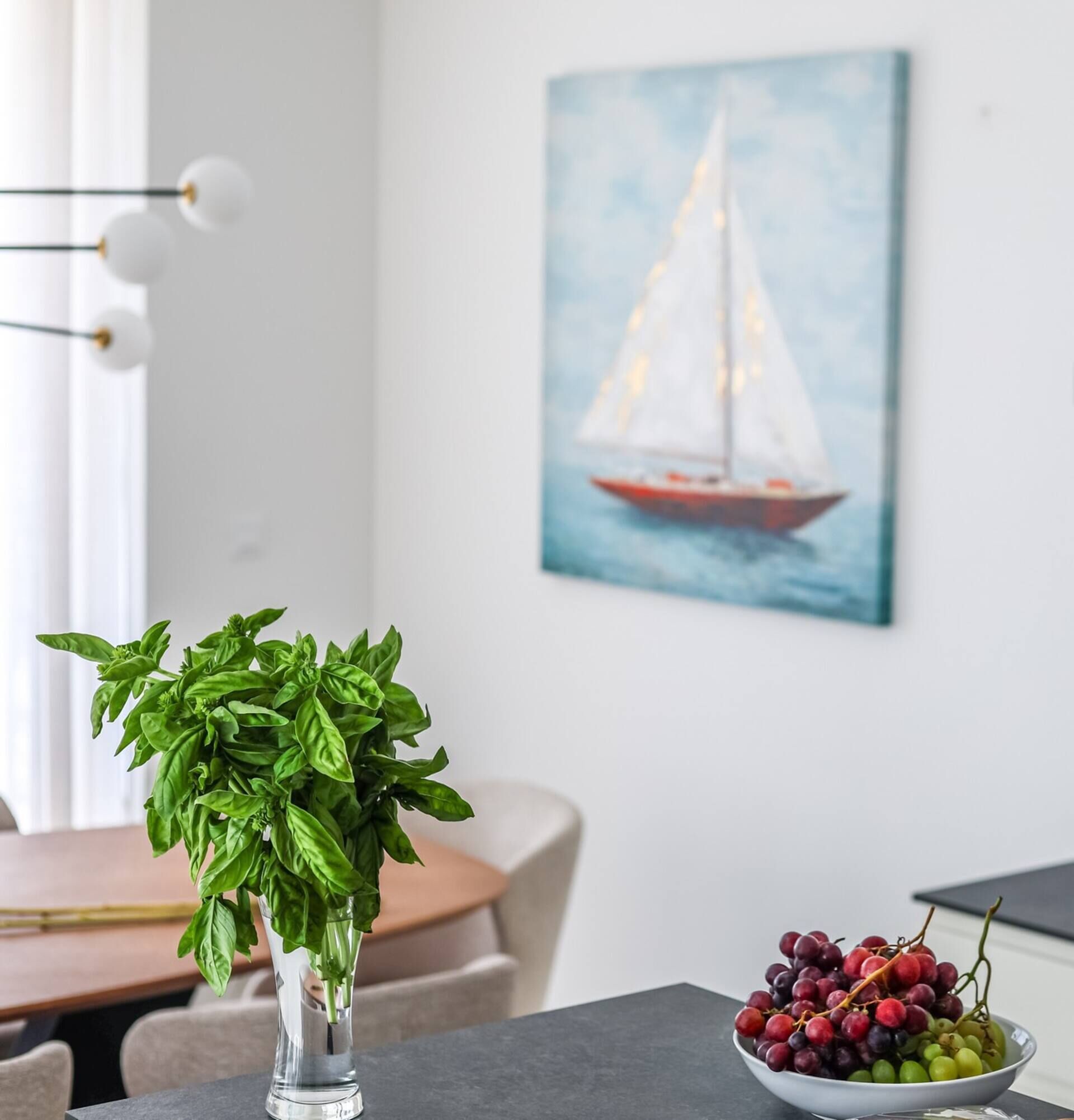 A dining table with a clear glass vase of basil, a bowl of mixed grapes, and a sailboat painting on the wall.