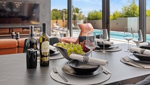 Mesa de comedor con botellas de vino, un cuenco de uvas y copas, con vista a una piscina y un área de estar al aire libre.