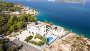 Un bâtiment moderne à deux étages, doté d’une piscine sur le toit et de panneaux solaires, qui offre une vue sur une zone côtière aux eaux bleues et limpides et sur une île située au loin.