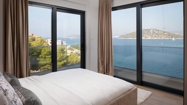 Un dormitorio con una cama grande, dos ventanas con cortinas y vistas al mar y al follaje.