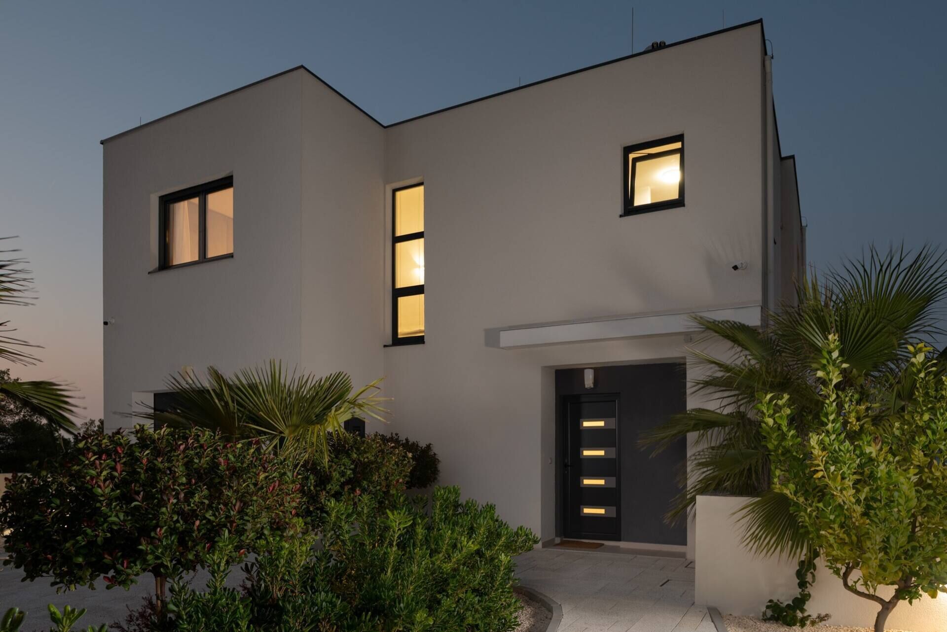 Una casa moderna de dos pisos con techo plano, ventanas amplias y una puerta de entrada oscura.