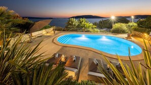Una piscina con vista al atardecer, rodeada de sillones de descanso y un muro de piedra.
