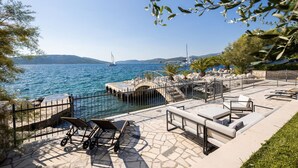 Un patio frente al mar con mobiliario exterior, una terraza de piedra y vistas al océano y veleros.