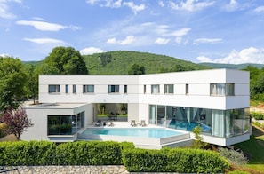 A modern two-story house with a swimming pool, large glass windows, and a view of green hills.