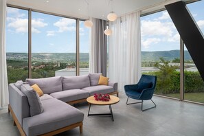 A modern living room with a grey sectional sofa, a blue armchair, and a round coffee table with flowers. Large windows offer a view of a green landscape and buildings.