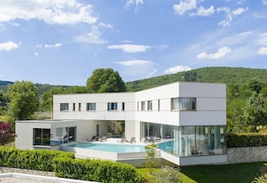 A modern two-story house with a swimming pool, large windows, and a view of a forested hill.