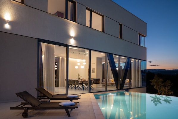 A modern house with a glass-enclosed dining area, a pool, and lounge chairs.