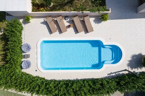 A rectangular swimming pool with a blue interior, surrounded by a white tiled deck. There are four lounge chairs and two round seating areas.