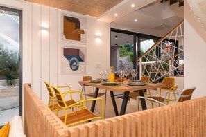 Un coin repas avec une table en bois dressée pour six personnes, entourée de chaises jaunes. Les murs sont ornés d’œuvres d’art abstraites, on y trouve un escalier avec une rampe aux motifs géométriques, et une vue sur la verdure à l’extérieur.