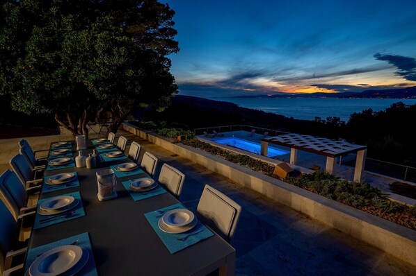 An outdoor dining area with a pool and a scenic view of the ocean at dusk.